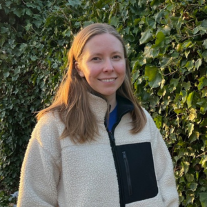A white woman with long blonde hair, wearing an off white fleece and stood in front of a green hedge.